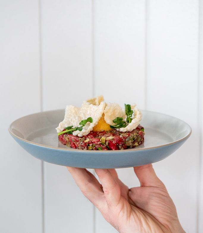 Hand holding Wagyu Tartare on a blue plate against a white backdrop